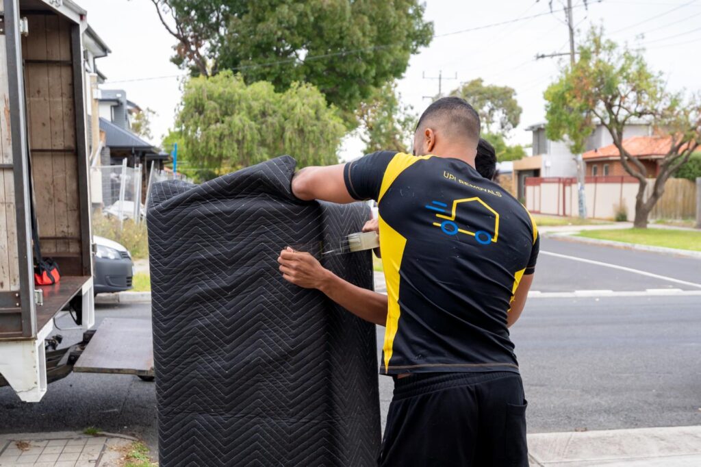 UP! Removals mover securing a furniture piece with moving blanket and tape beside a truck on a residential street