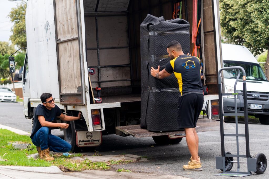 UP! Removals movers loading a large item wrapped in a moving blanket into a truck using the tail lift, with a hand trolley on site